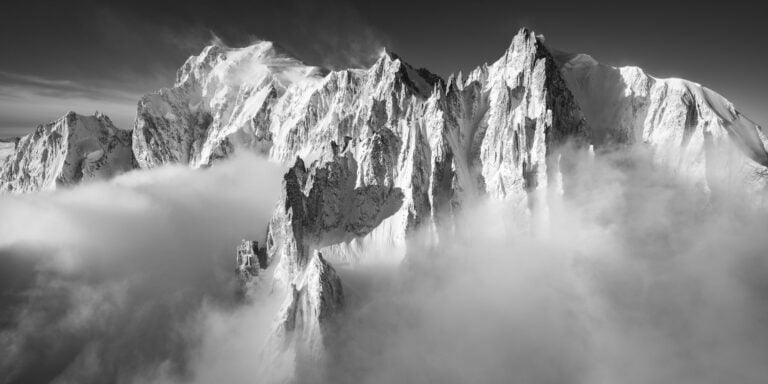 hotographie noir et blanc du massif du Mont Blanc avec les Aiguilles du Diable, le Grand Capucin et le Mont Maudit illuminés par la lumière matinale après la tempête.