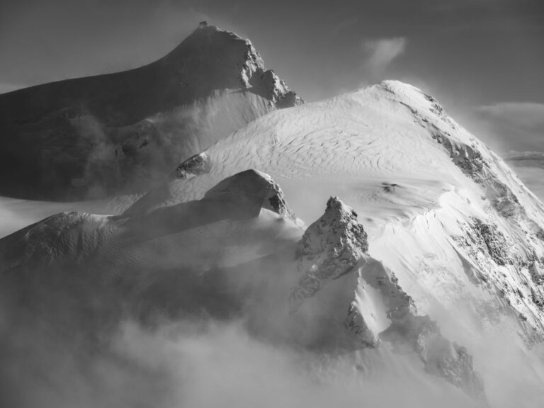 Schwarz-Weiß-Fotografie der Gipfel des Monte-Rosa-Massivs, mit der Parrotspitze und der Gnifetti-Spitze, die aus dem Nebel auftauchen und vom Wind geformt werden.